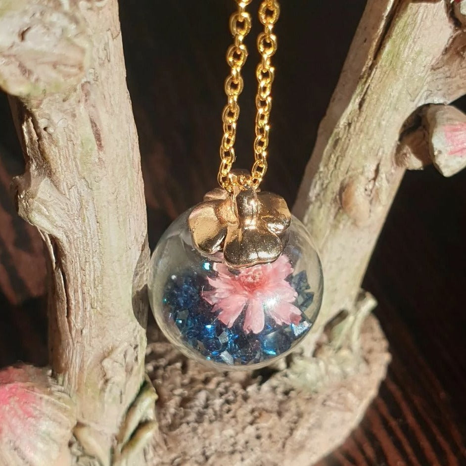 This Blue Jewel Globe Necklace is the perfect way to show off your unique style! Crafted with an elegant glass globe pendant filled with dark blue jewel stones and a cute pink flower, this necklace will add a sparkle of sophistication to any outfit. With a golden chain for added flair, you'll look beautiful and timeless!