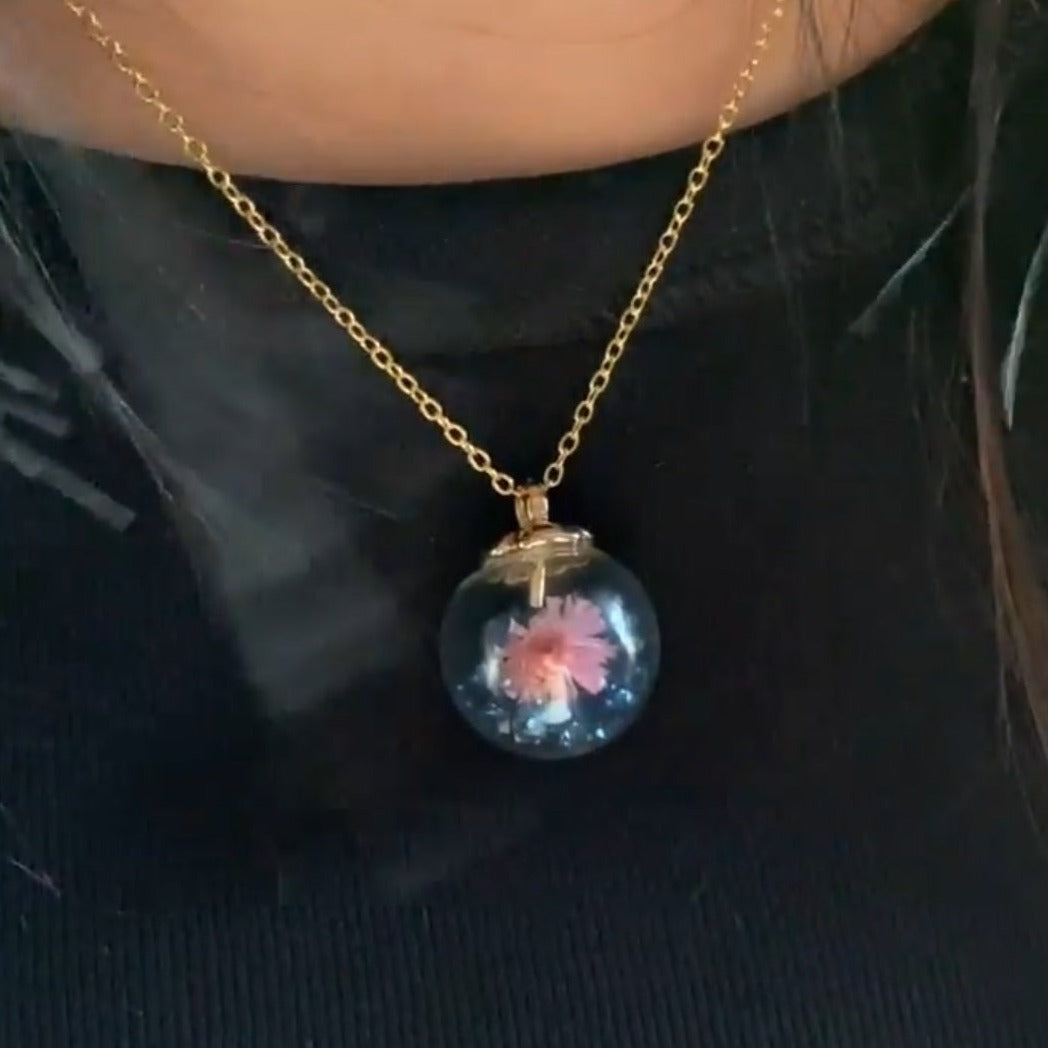 This Blue Jewel Globe Necklace is the perfect way to show off your unique style! Crafted with an elegant glass globe pendant filled with dark blue jewel stones and a cute pink flower, this necklace will add a sparkle of sophistication to any outfit. With a golden chain for added flair, you'll look beautiful and timeless!